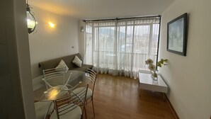 A dining area with a glass table and chairs, a sofa with cushions, a window with sheer curtains, and a framed picture on the wall.