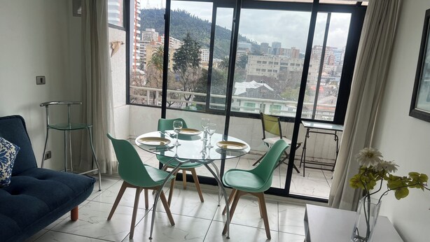 A modern living room with a dining area, large windows with a city view, a blue sofa, and a dining table set for four.