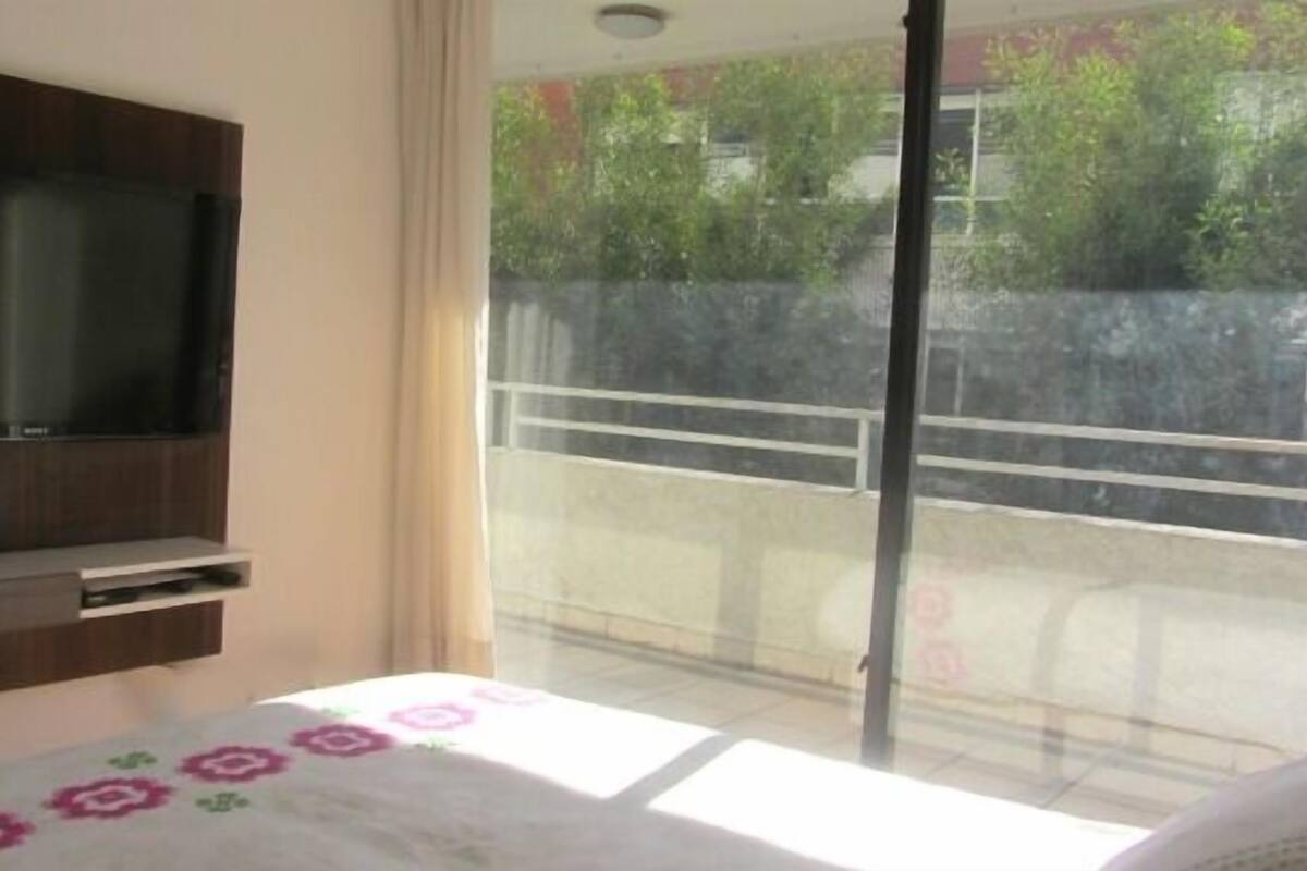 A bedroom with a TV mounted on the wall, a bed with a floral bedspread, and a balcony with a view of greenery.