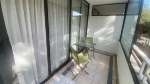 A balcony with a glass table and chairs, a view of the street, and a large window with curtains.