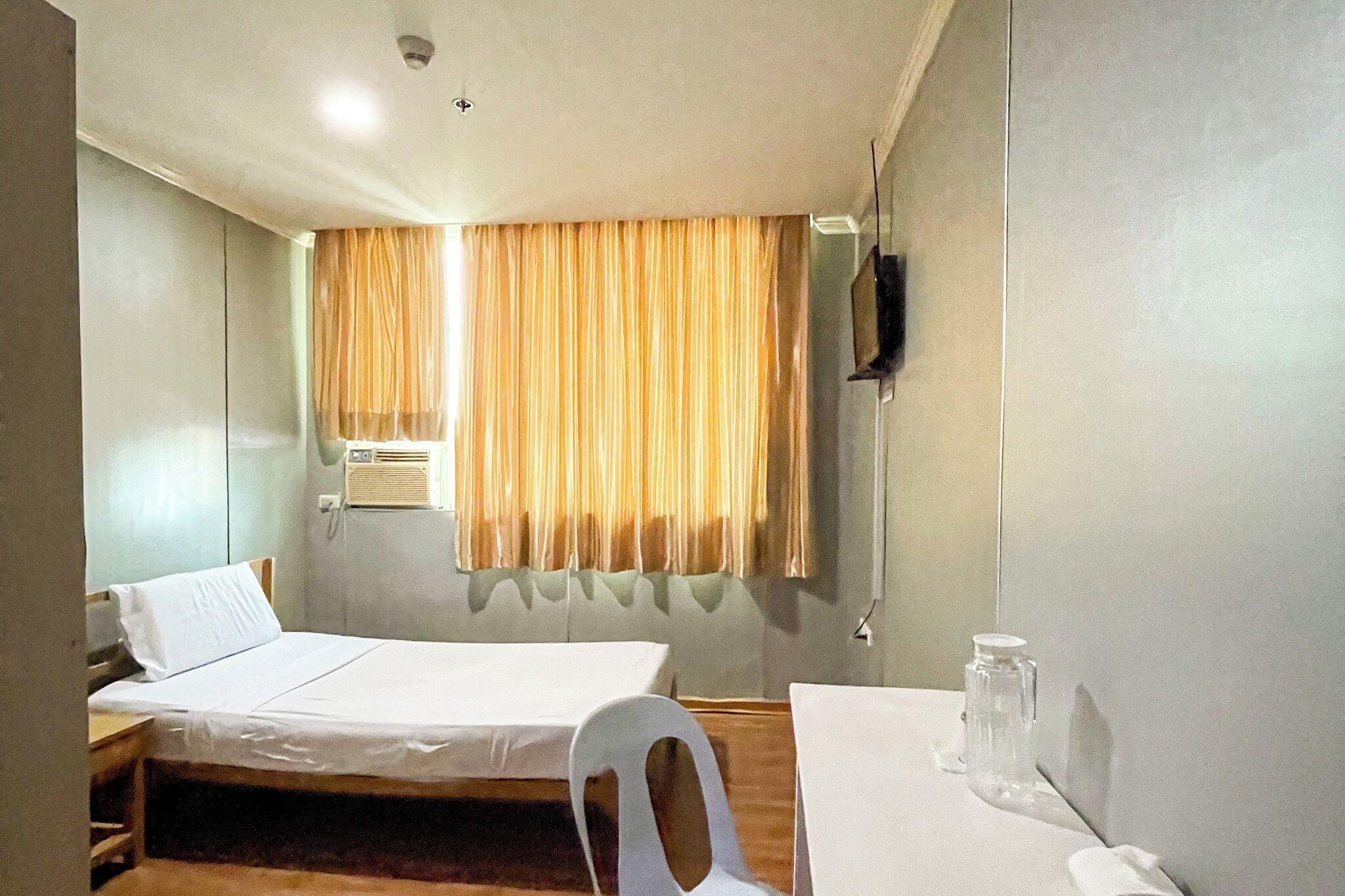 A single bed with a white mattress and pillow, a small table with a chair, an air conditioning unit, and a window with curtains.