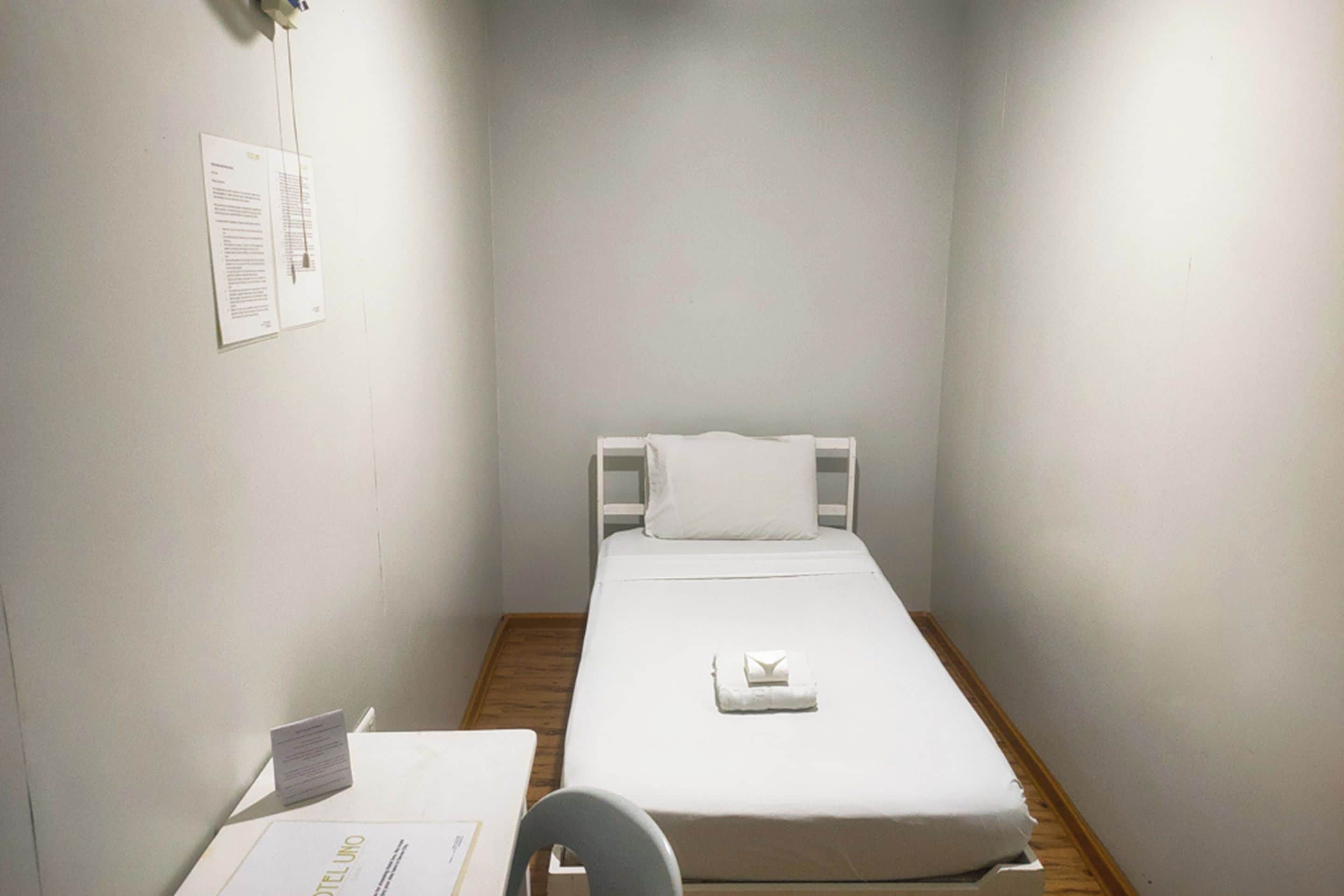 A small, single-bed room with a wooden floor, white walls, and a white bedspread. There is a desk with a chair and a small table with a sign on it.