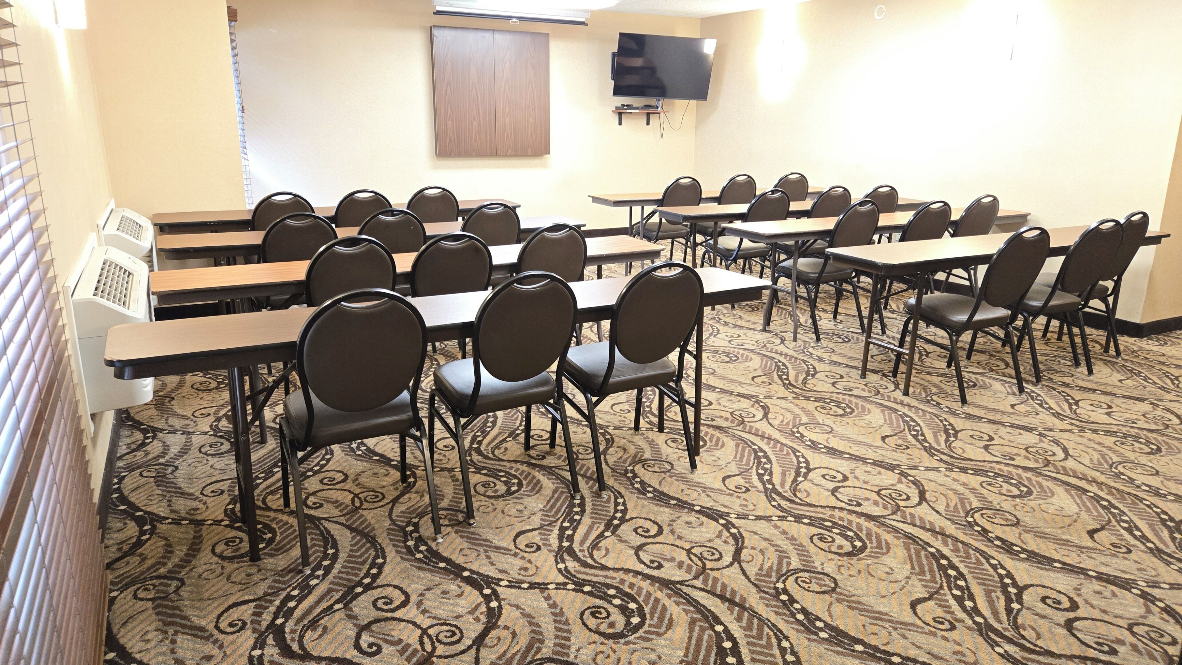 A conference room with multiple tables and chairs, a carpeted floor, a television on the wall, and a window with blinds.