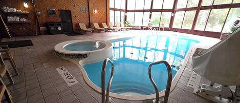 An indoor swimming pool with a hot tub, lounge chairs, and a view of the outdoors.
