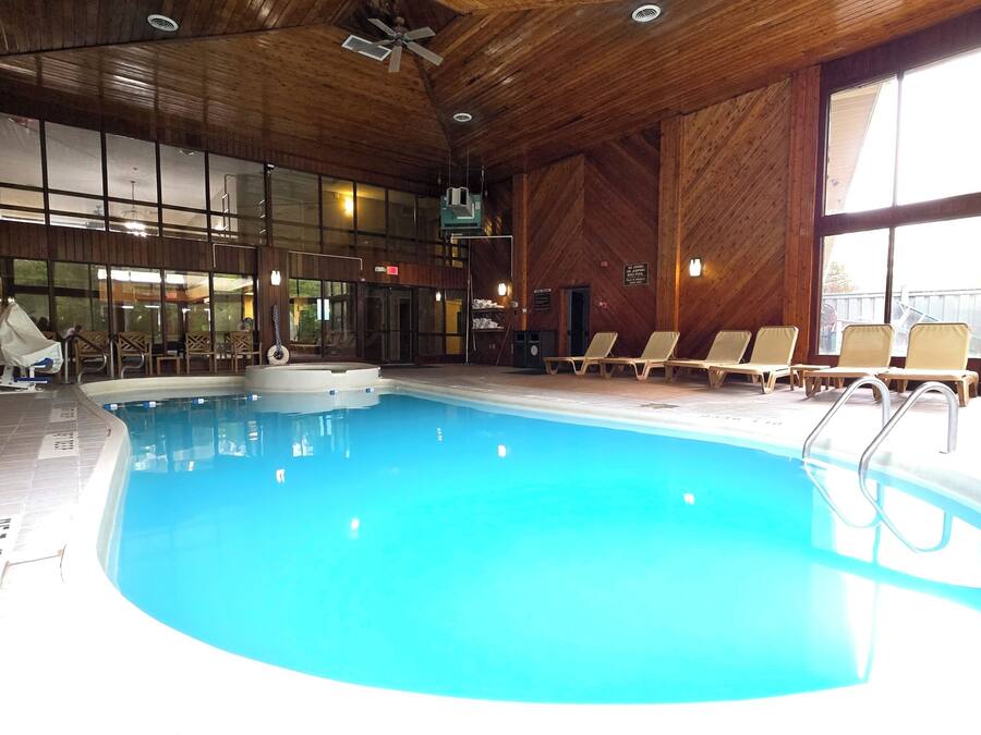 Una piscina interior con techo de madera, ventanas iluminadas por el sol y sillas de descanso.