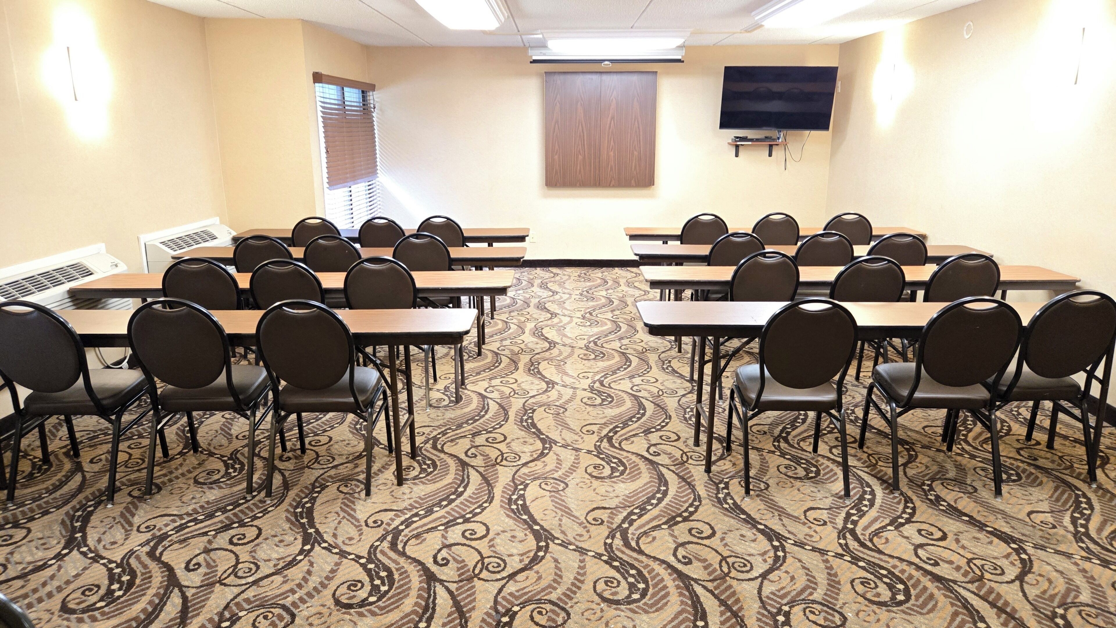A conference room with a patterned carpet, rows of tables and chairs, a television on the wall, and a window with blinds.
