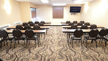 A conference room with a patterned carpet, rows of tables and chairs, a television on the wall, and a window with blinds.