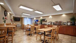 Un comedor de hotel con mesas y sillas de madera, una barra de cocina con electrodomésticos y un televisor en la pared.