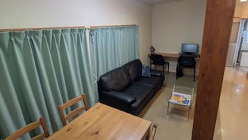 A room with a wooden table and chairs, a black leather sofa, a small glass table with a magazine, a computer on a desk, and a green curtain.