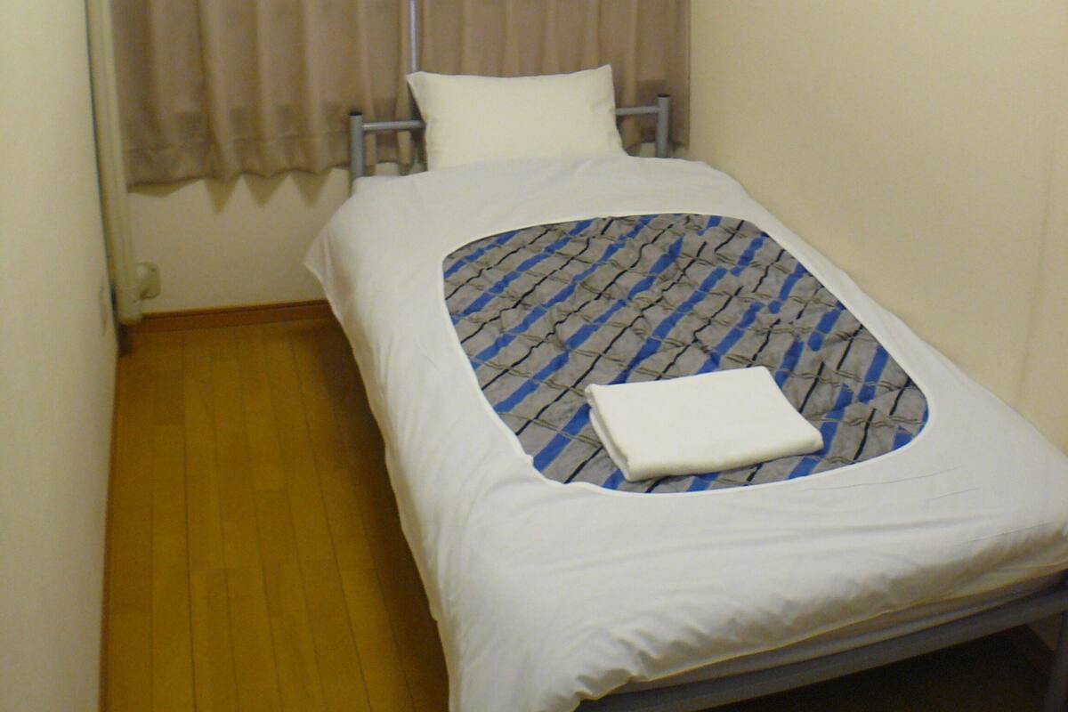 A small, single-bed room with a wooden floor, a white bedspread, a headboard with a curtain, and a wall-mounted air conditioner.