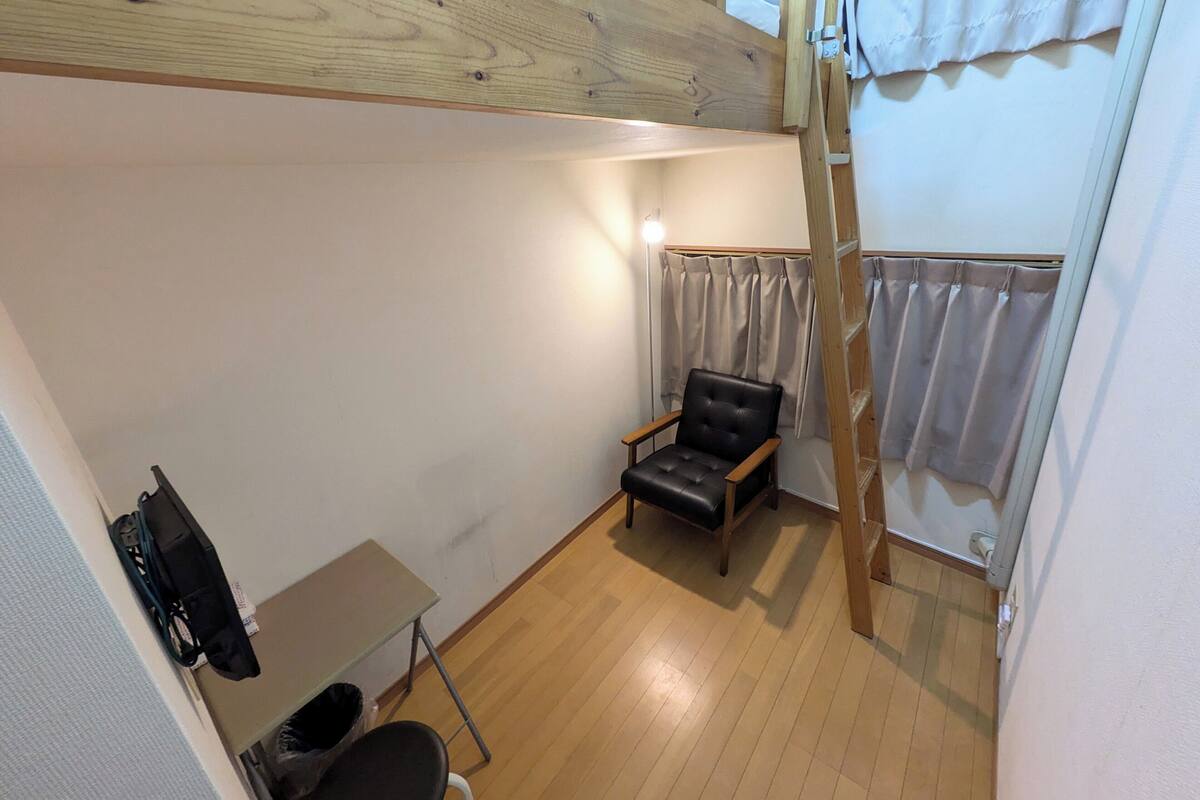 A room with a loft bed, a chair, a small table, and a window with curtains.