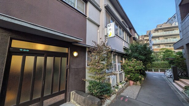 Un estrecho callejón con un edificio tradicional japonés, puerta corredera y un pequeño jardín con plantas en macetas.