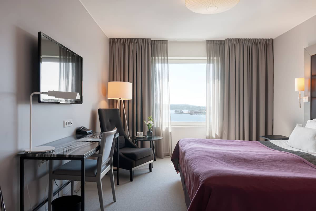 A hotel room with a bed, desk, chair, and a large window with curtains.