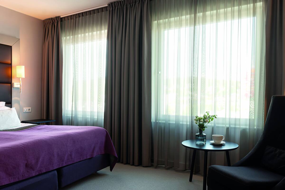 A hotel room with a bed, a chair, a small table with a vase and cup, and sheer curtains.
