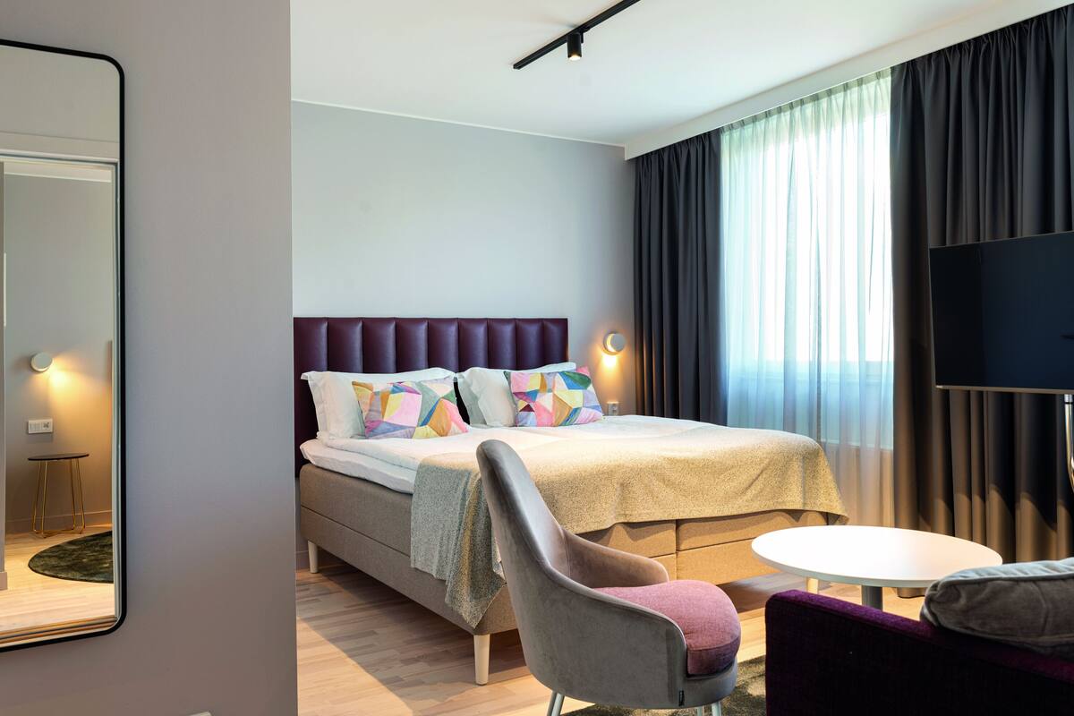 A modern hotel room with a large bed, a flat-screen TV, a round table, and a comfortable chair.