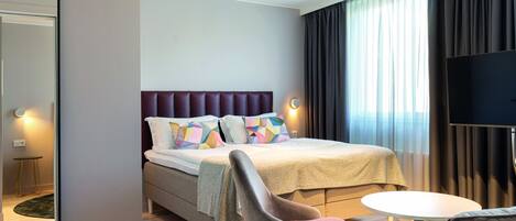 A modern hotel room with a large bed, a flat-screen TV, a round table, and a comfortable chair.
