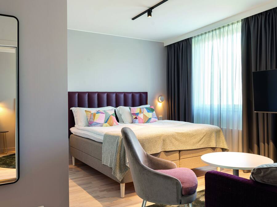 Una habitación de hotel moderna con una cama grande, un televisor de pantalla plana, una mesa redonda y una silla cómoda.