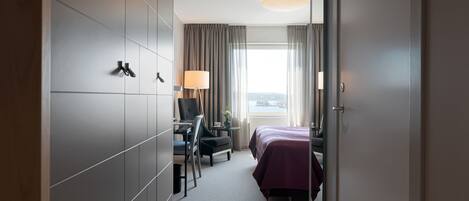 Ein Hotelzimmer mit Bett, Schreibtisch und Stuhl. Ein Fenster mit Vorhängen, eine Lampe und Blick auf die Stadt.