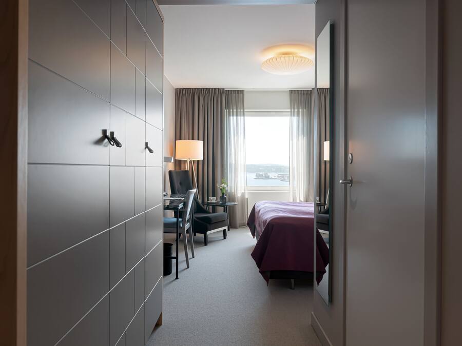 Habitación de hotel con cama, escritorio y silla. Tiene una ventana con cortinas, una lámpara y vistas a la ciudad.