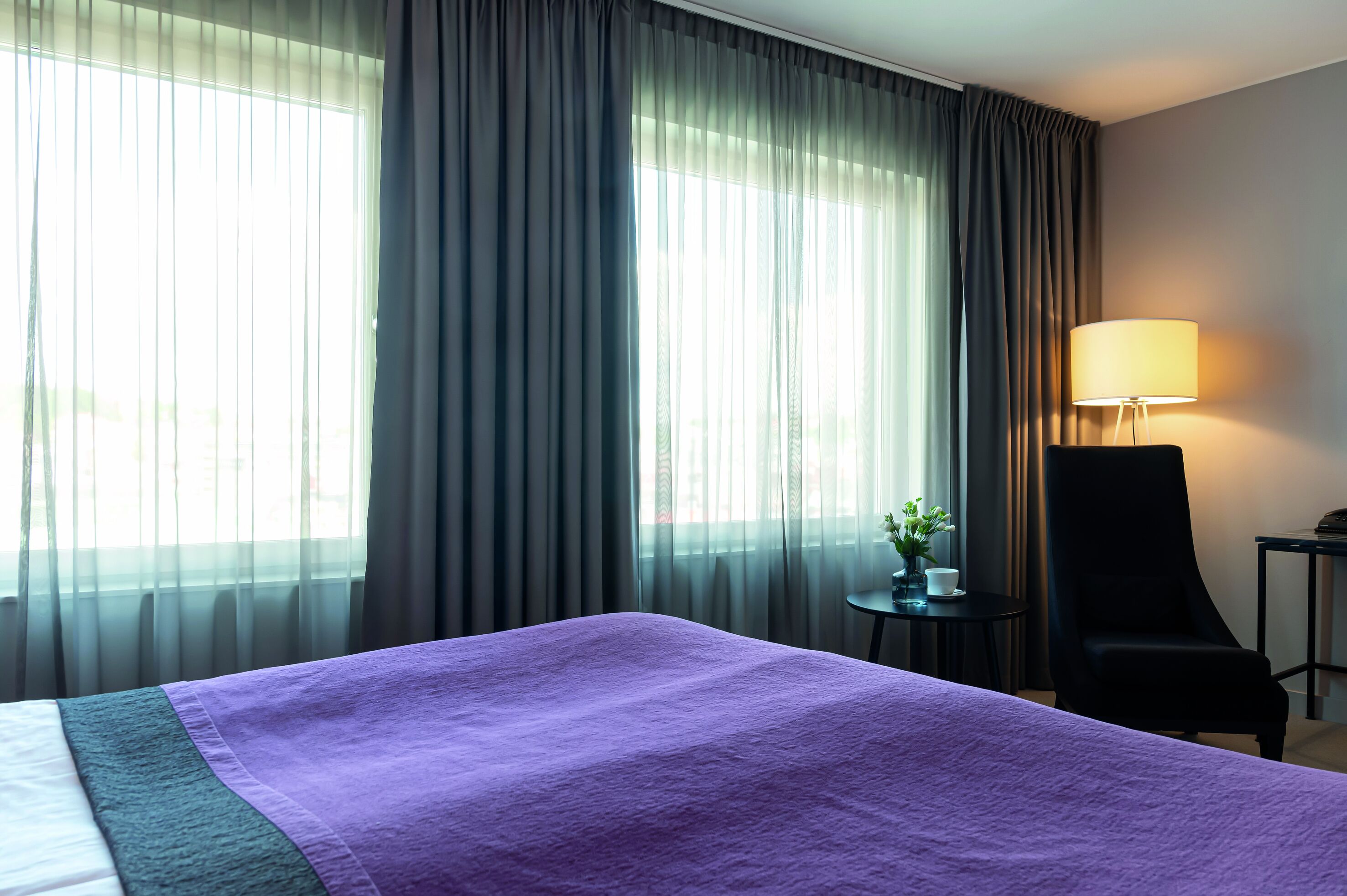 A hotel room with a large bed, a chair, a small table with a lamp, and sheer curtains on the windows.