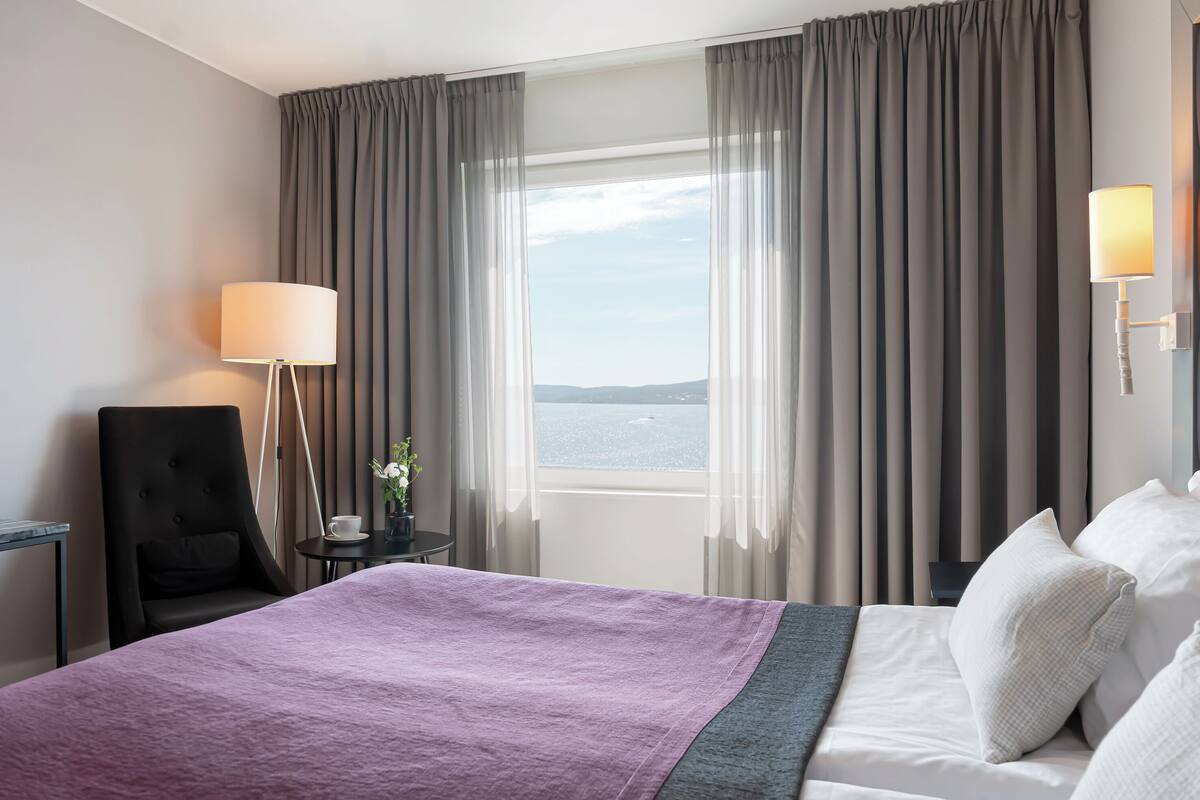 A hotel room with a large bed, a chair, a small table with a lamp, and a view of the sea through sheer curtains.