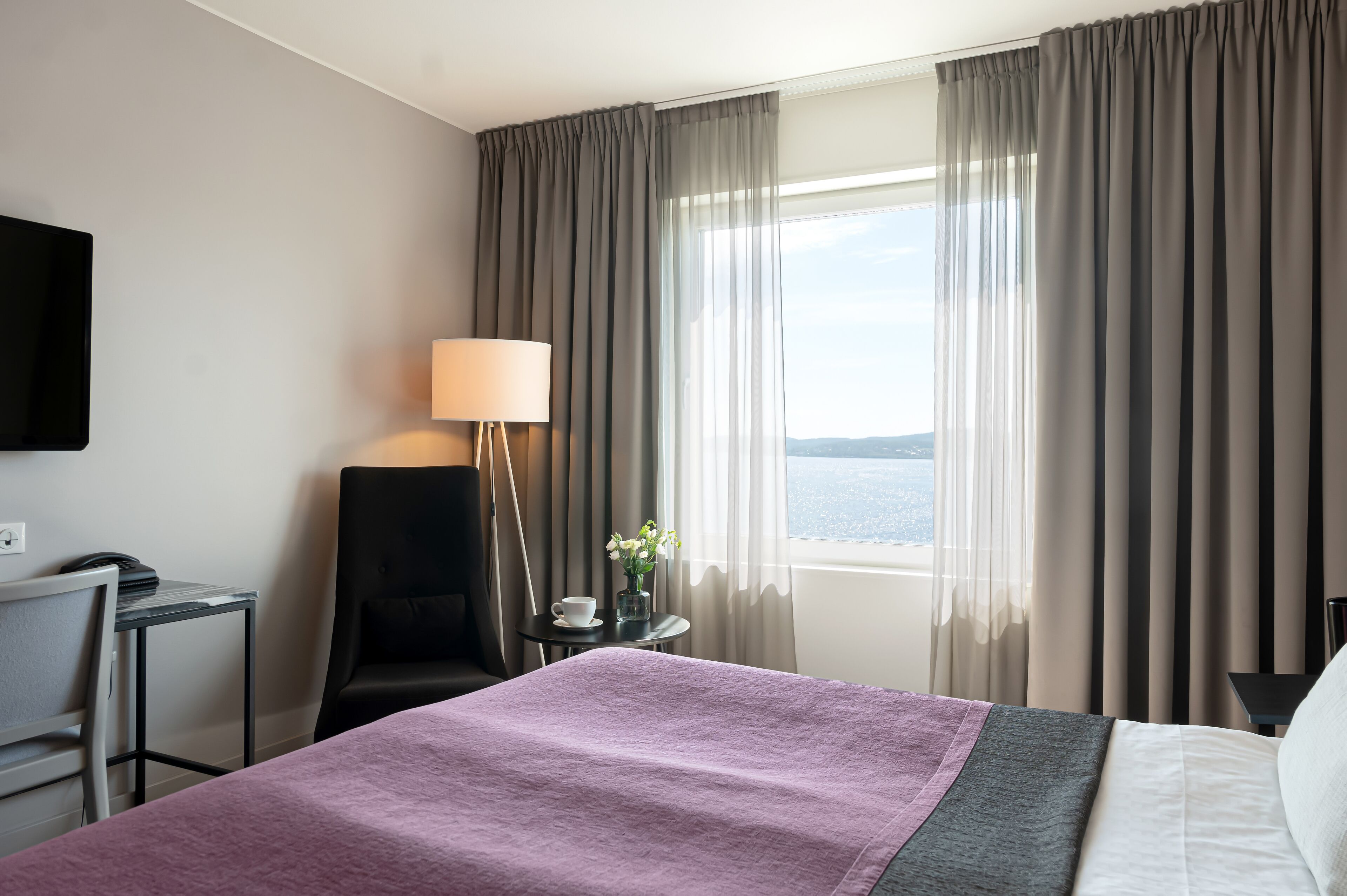 Ett hotellrum med en säng, ett skrivbord, en stol, en lampa och utsikt över havet.