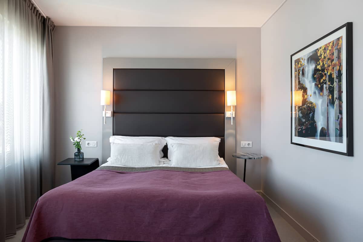 A modern hotel room with a large bed, two bedside lamps, a nightstand with a vase, and a framed artwork on the wall.