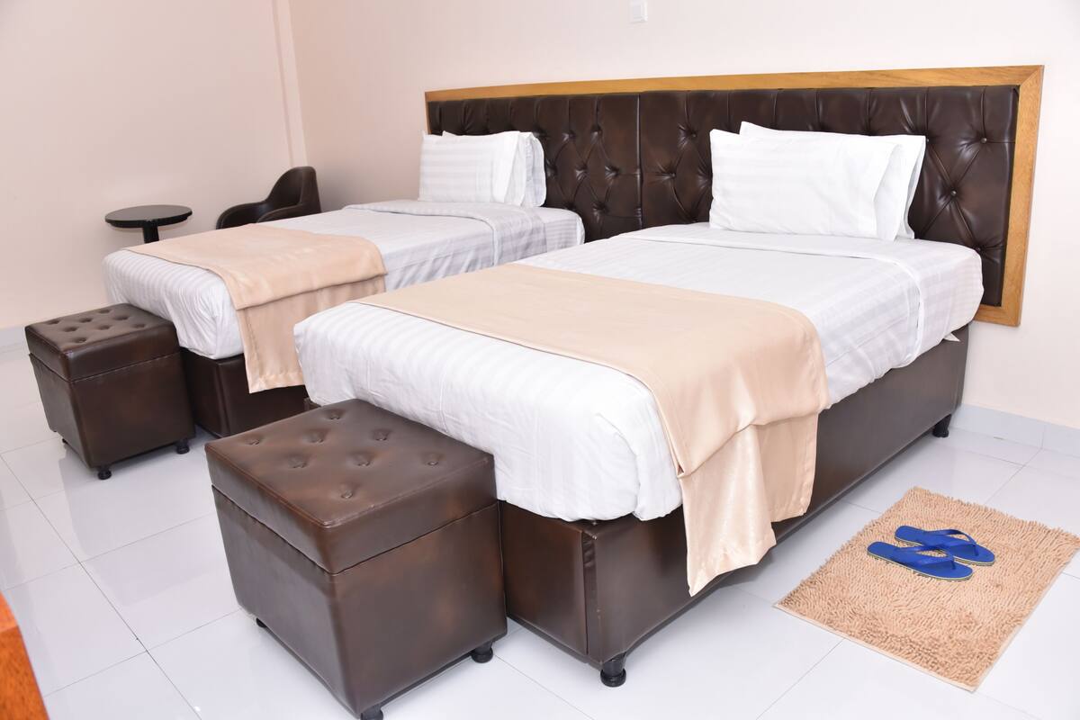 Une chambre d’hôtel avec deux lits, une petite table, une chaise, un pouf marron et une paire de chaussons posés sur un tapis.