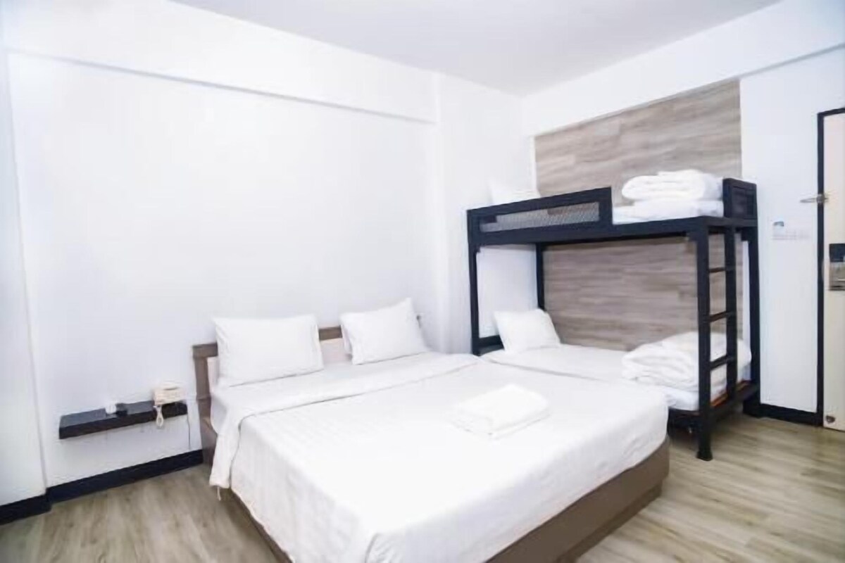 A hotel room with a bunk bed, a single bed, white bedding, a wooden floor, and a door with a towel rack.
