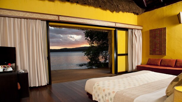 Een slaapkamer met een bed, een televisie en uitzicht op een meer bij zonsondergang.