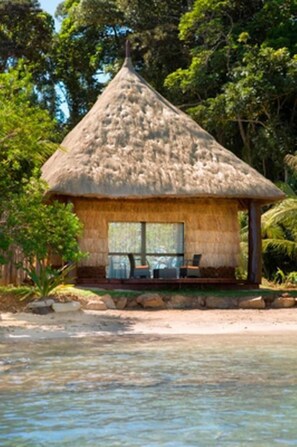 Une hutte au toit de chaume, dont l’intérieur est visible, située au bord d’une étendue d’eau calme.