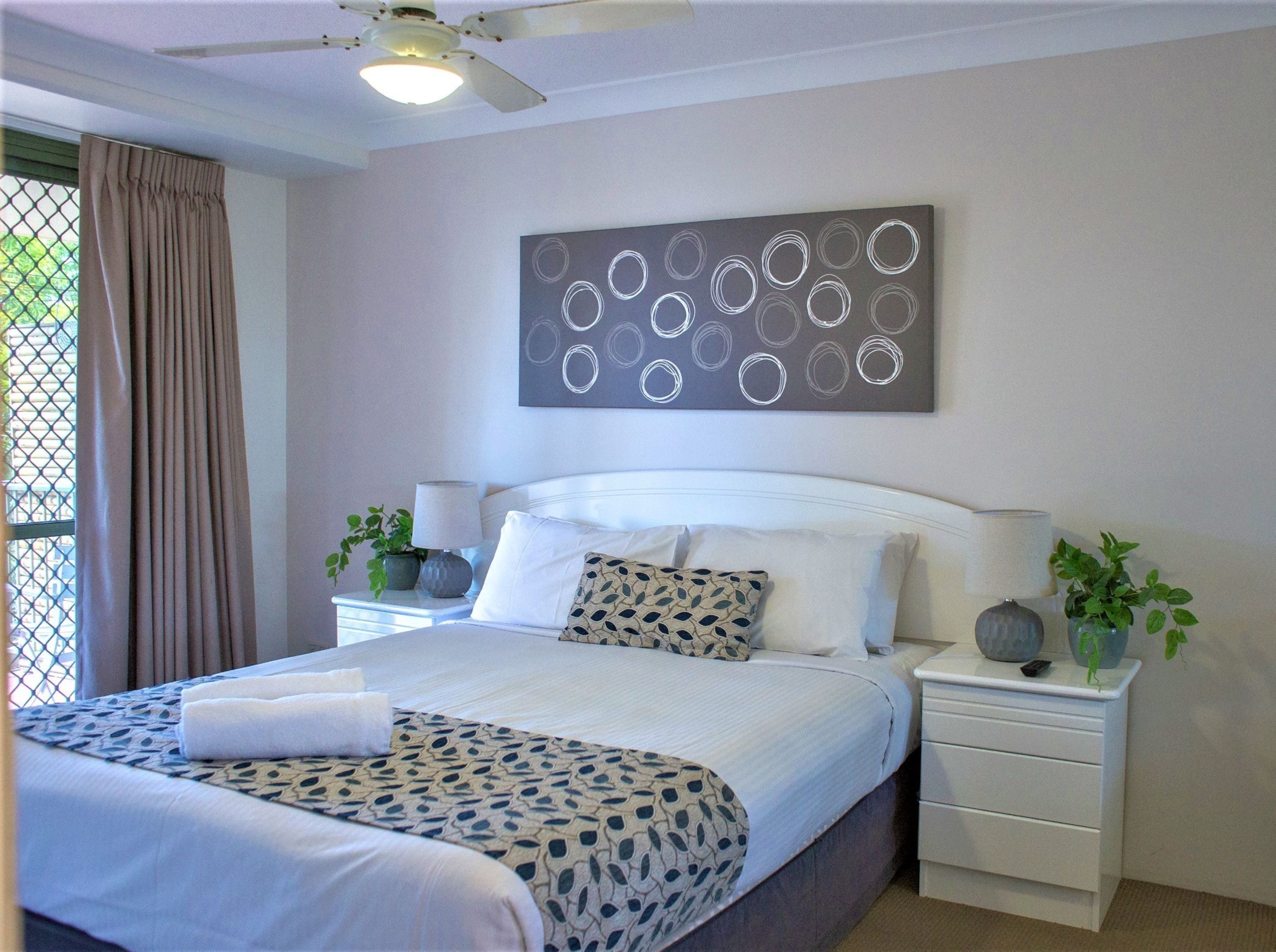 A neatly made bed with a patterned bedspread, a white headboard, and a bedside table with a lamp and a plant.