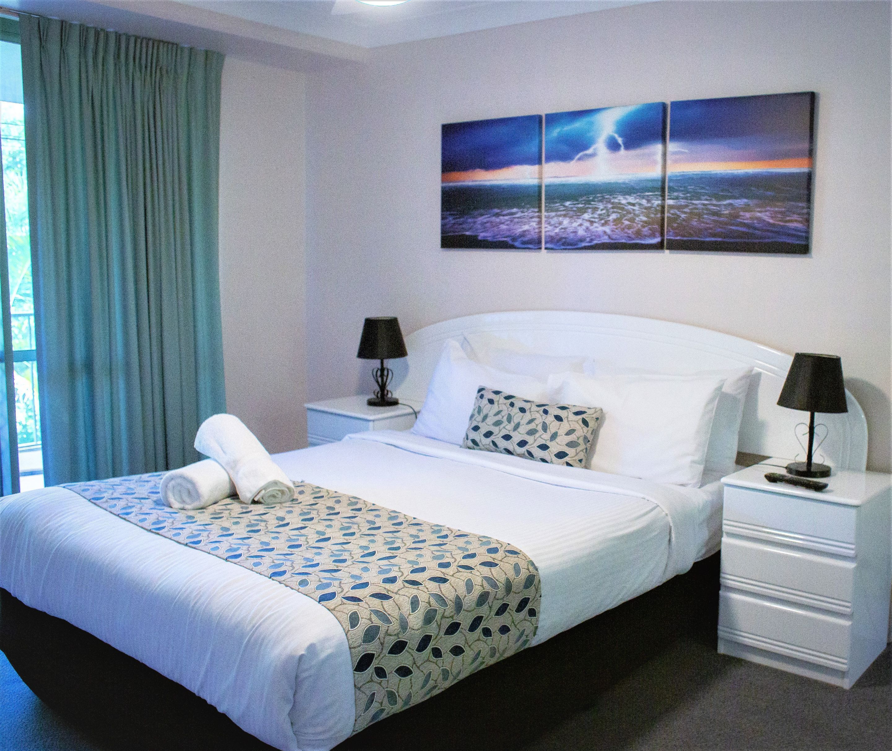 A neatly made bed with a patterned bedspread, two bedside lamps, a nightstand, and a wall-mounted painting.