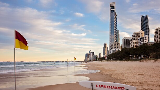 Una playa con un puesto de salvavidas, una tabla de surf, una bandera y un horizonte urbano al fondo.