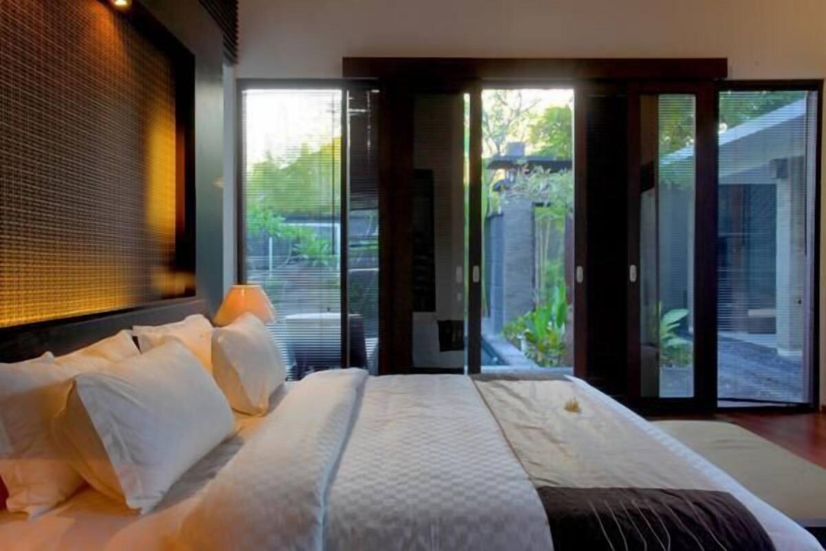 A modern hotel room with a large bed, a view of the garden through sliding glass doors, and a wall with a textured pattern.