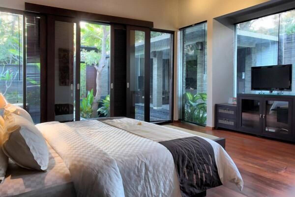 A bedroom with a large bed, sliding glass doors leading to a garden, a television on a cabinet, and a dark-colored wardrobe.
