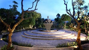 Une fontaine circulaire en pierre, ornée de statues, entourée d’arbres et bordée d’un chemin pavé.