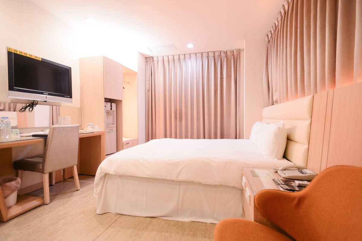 A hotel room with a bed, a desk with a computer, a TV, and a window with curtains.