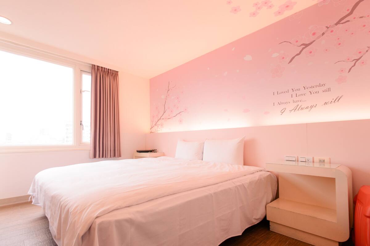 A bedroom with a large bed, a bedside table, a nightstand, a wall-mounted shelf, and a window with curtains.
