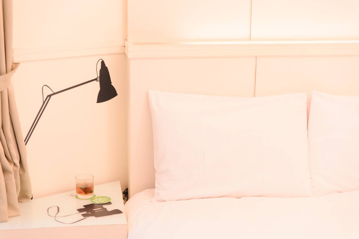A neatly made bed with white pillows and a bedside table with a lamp, a glass of water, and a pair of earphones.