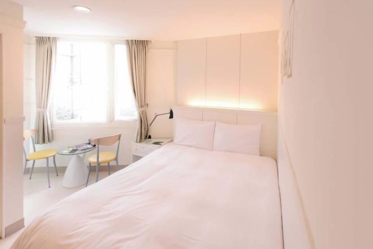 A hotel room with a bed, a small table, two chairs, a window with curtains, and a wall-mounted lamp.