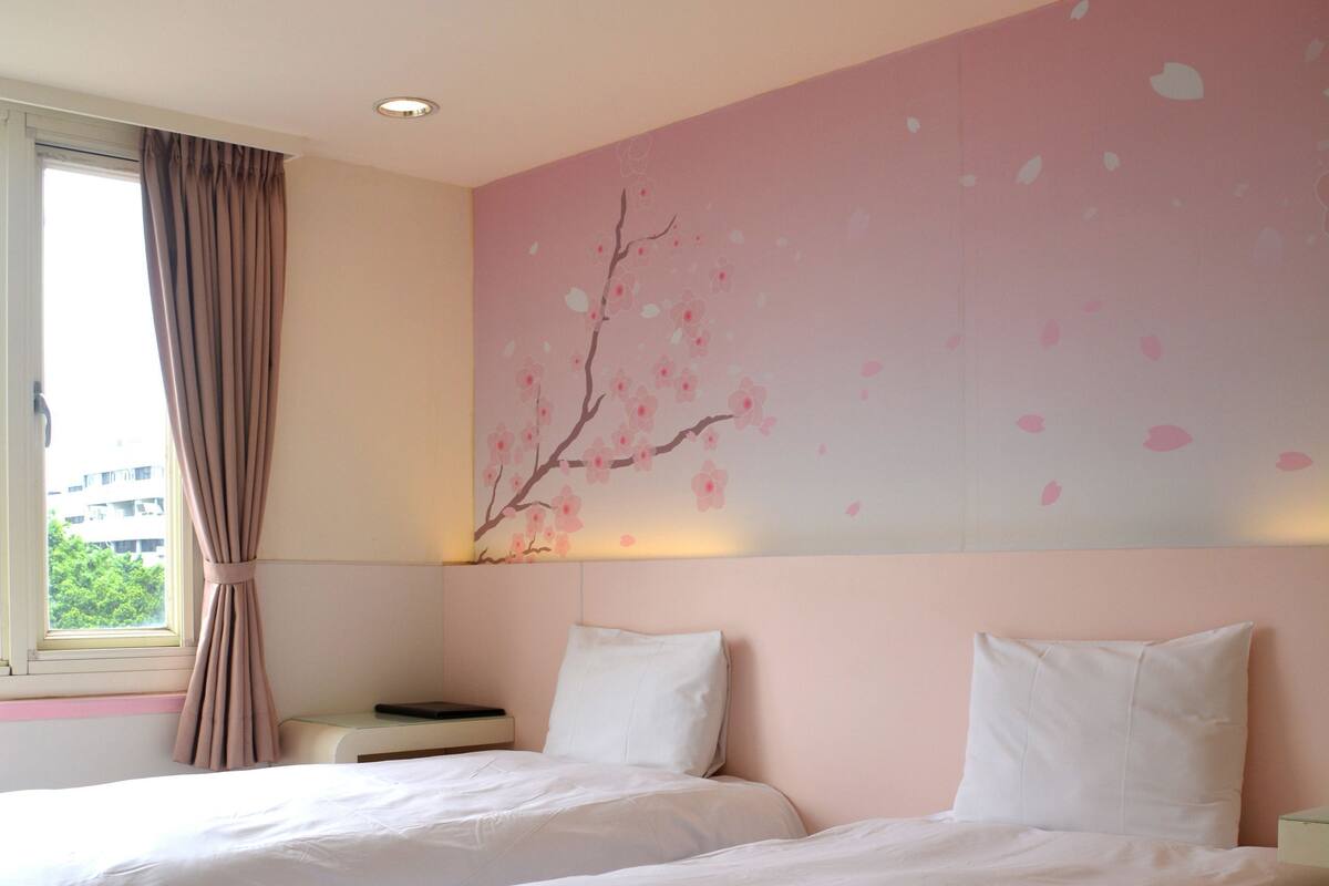 A hotel room with two beds, a window with curtains, a wall with a cherry blossom mural, and a ceiling vent.