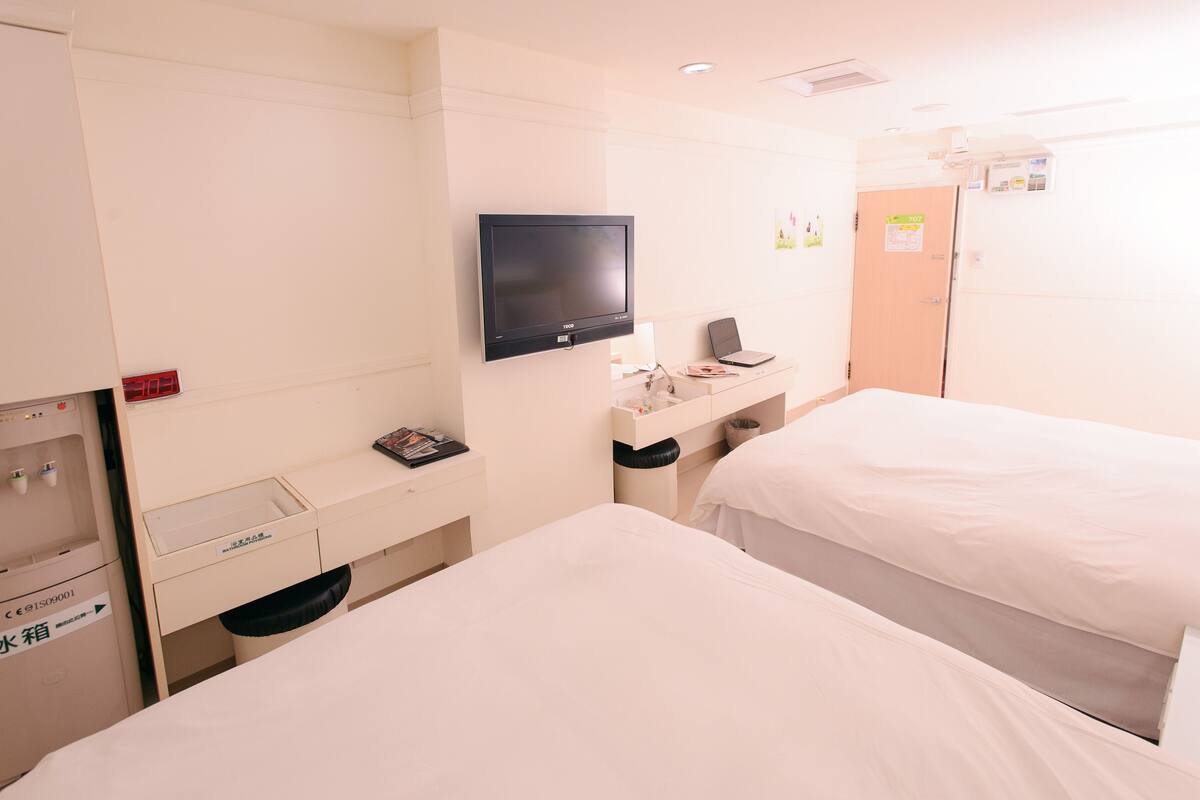A hotel room with two beds, a TV, a desk with a laptop, a trash bin, and a water dispenser.