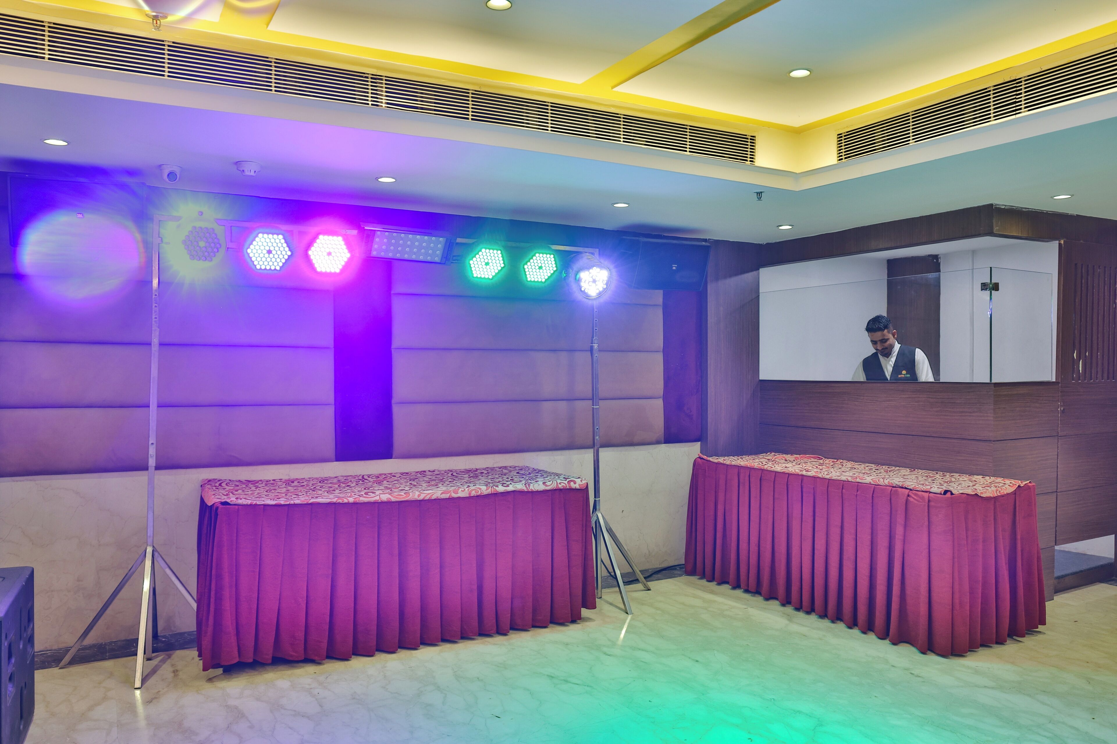 A conference room with two tables covered in red tablecloths, a ceiling with recessed lighting, and a large screen on the wall.