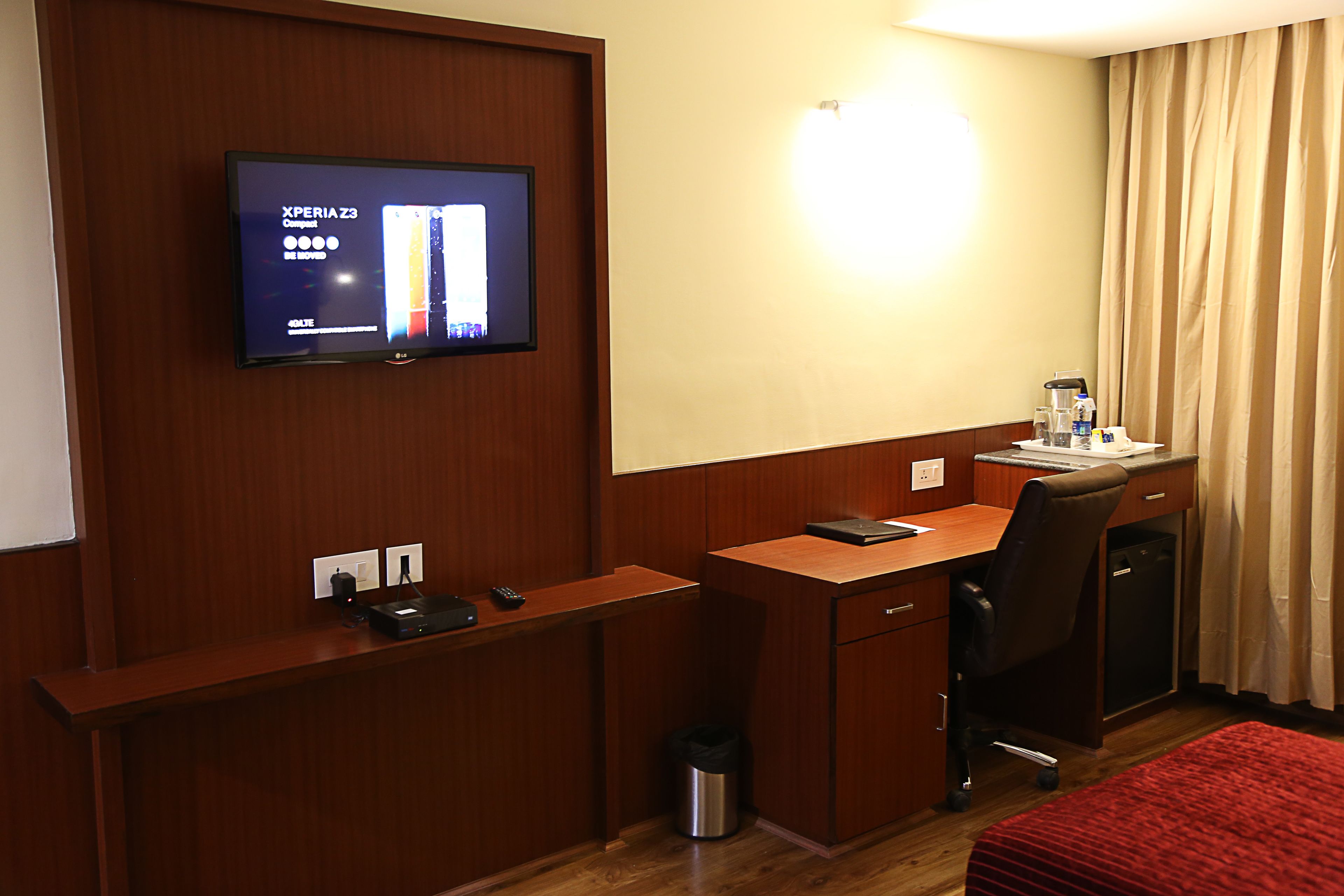 Quarto de hotel com secretária de madeira, televisão fixada na parede, um cesto de lixo pequeno e um edredom vermelho.