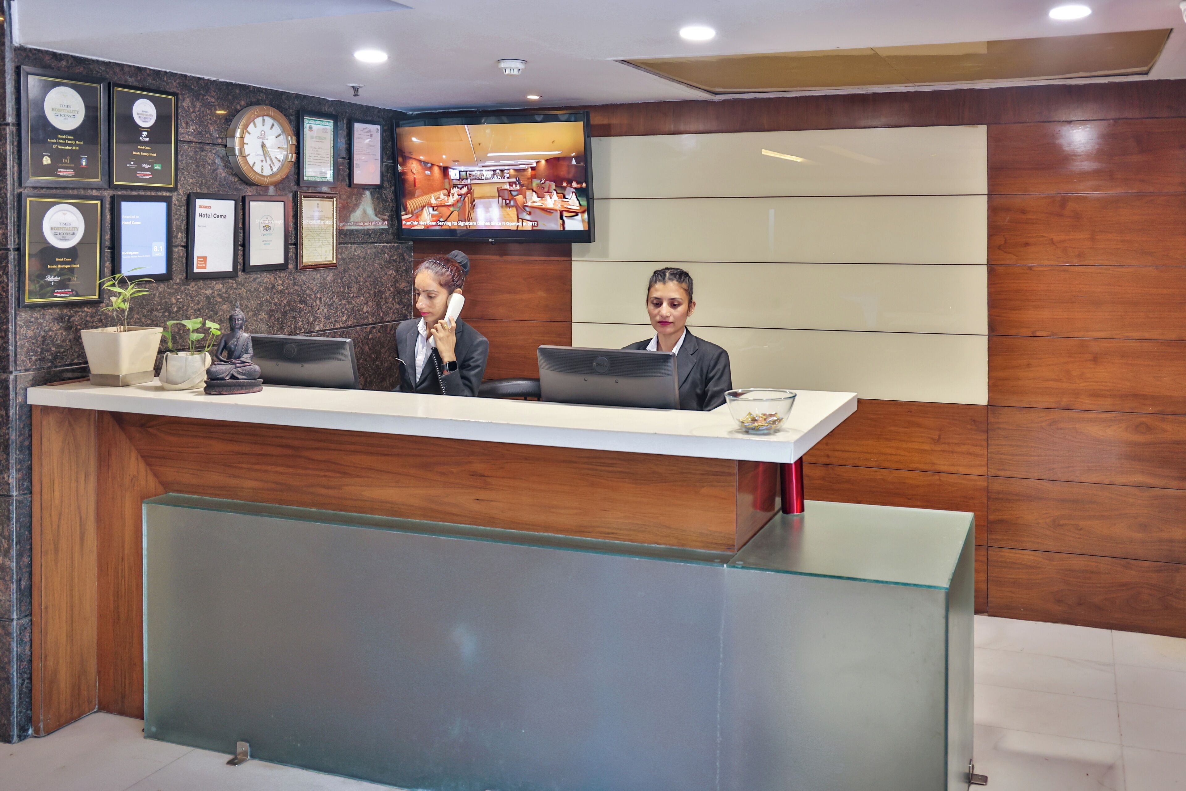 A reception desk with two staff members, a clock, framed certificates, and a television displaying a restaurant scene.