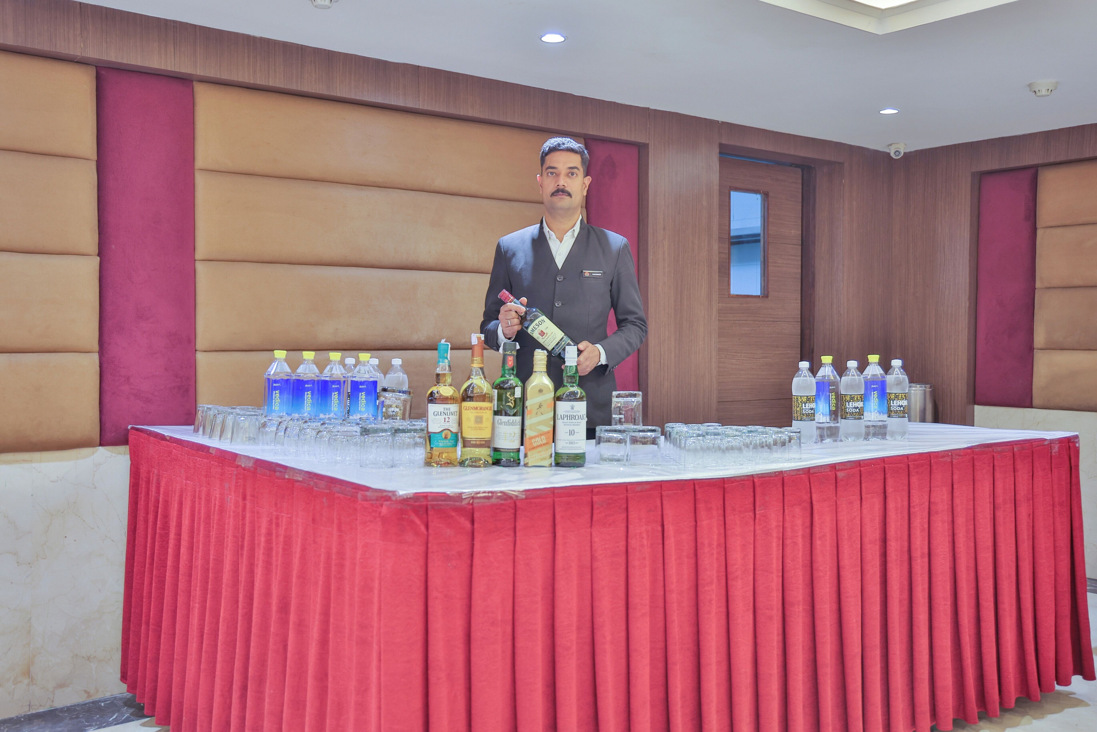 A man in a suit pouring a drink from a bottle onto a table with various liquor bottles and glasses.