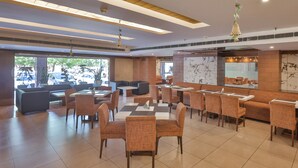 A modern restaurant with high tables and cushioned seating, large windows, and a view of the street. - Hotel Cama (Mohali)