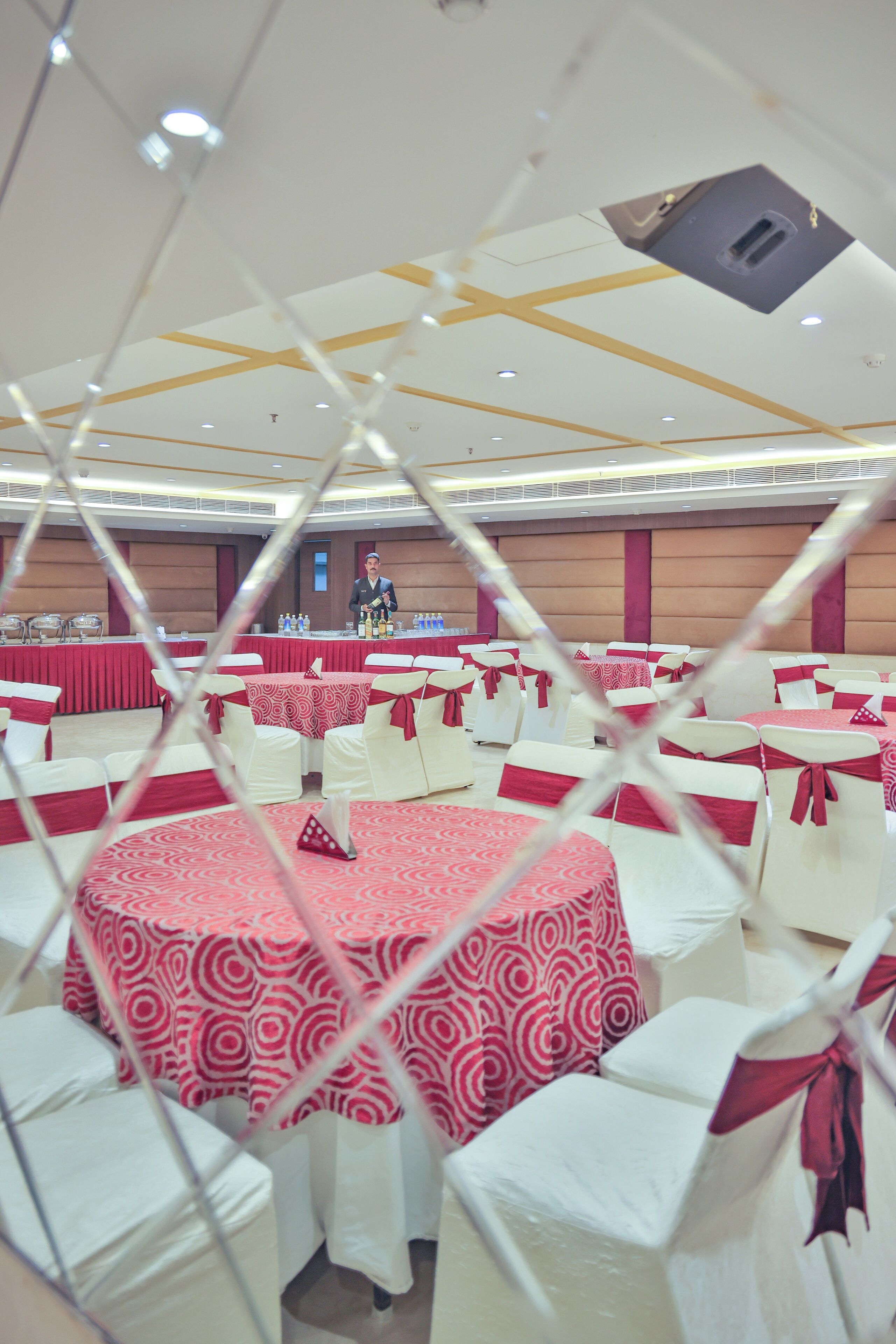 A banquet hall with round tables covered in red and white tablecloths, white chairs with red sashes, and a ceiling with recessed lighting.