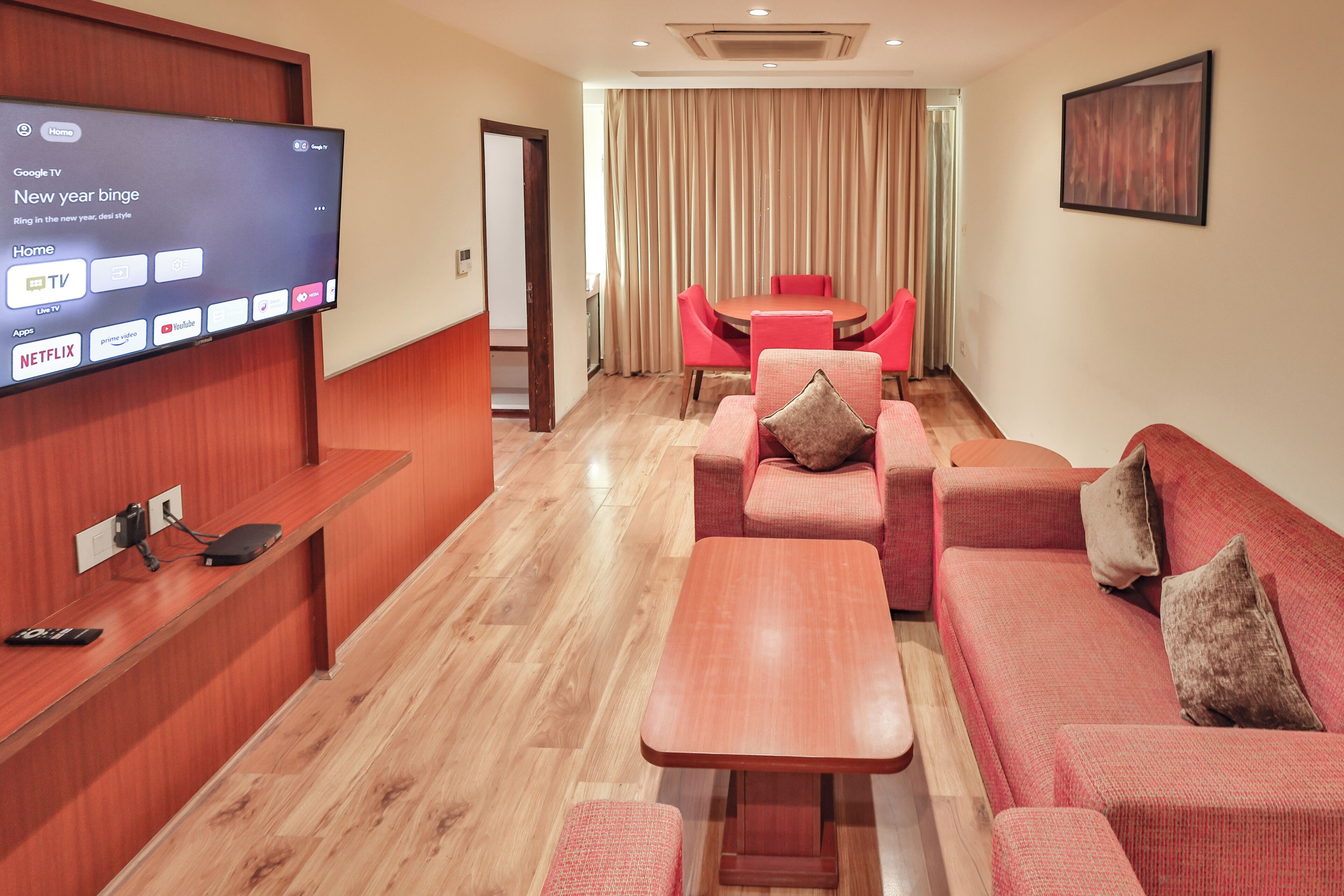 A hotel room with a flat-screen TV mounted on the wall, a wooden reception desk, a seating area with a sofa and armchairs, a dining table with chairs, and a large window with curtains.